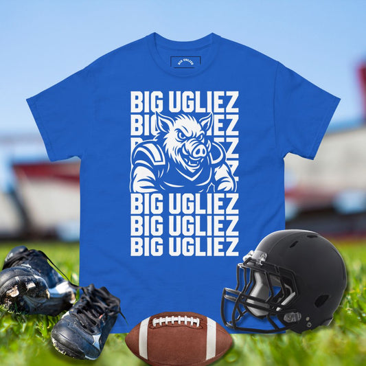 Blue t-shirt with 'BIG UGLIEZ' text and pig graphic, surrounded by football gear on grass.