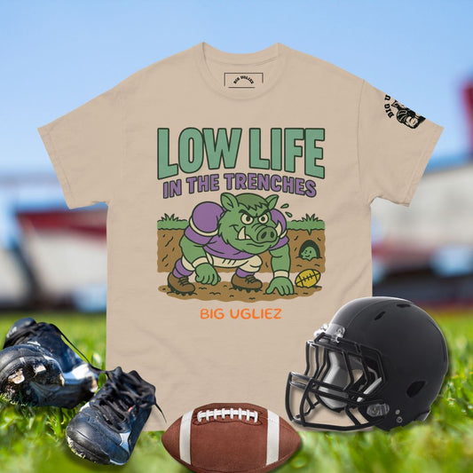 Beige t-shirt with graphic design of a piglet and text, placed on grass with football helmet, shoes, and ball.