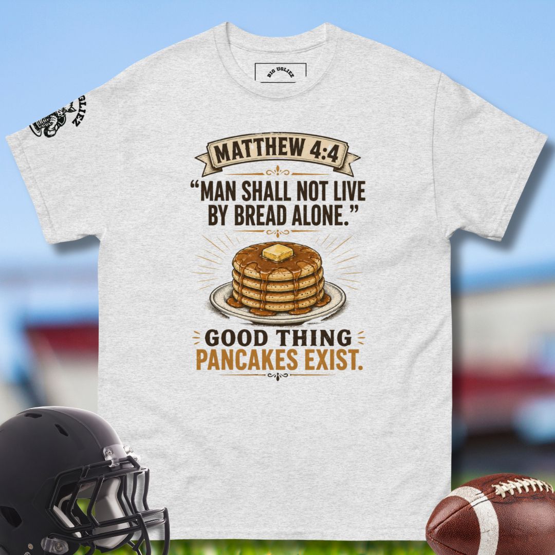 Gray t-shirt with a humorous design about pancakes, displayed with a helmet and football on a blurred sports field background.