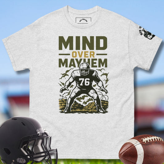 Gray t-shirt with football player graphic and 'Mind Over Mayhem' text, helmet, and football on a sports field background.
