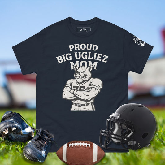 Black t-shirt with 'Proud Big Ugliez Mom' graphic, football, helmet, and shoes on grass.