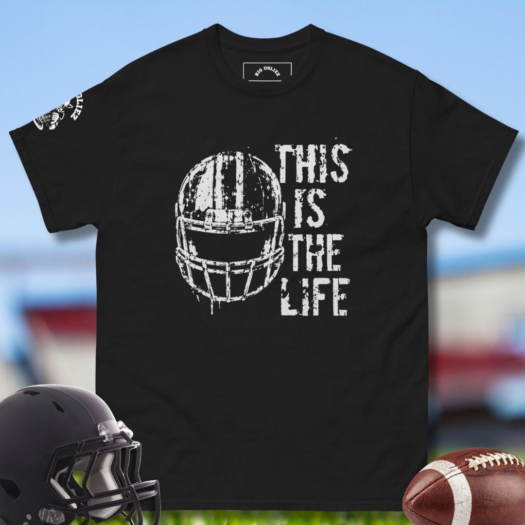Black t-shirt with football helmet graphic and 'This is the Life' text, displayed with a helmet and football on a sports field background.