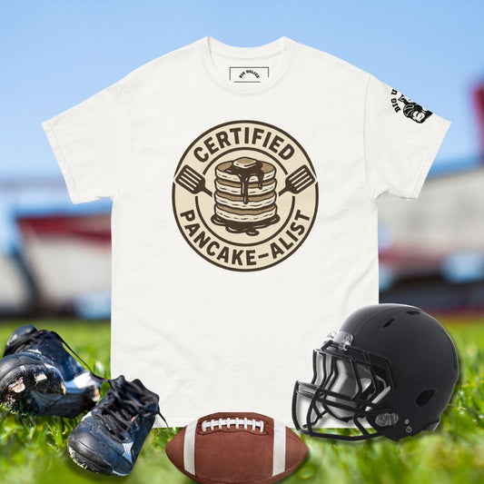 White t-shirt with 'Certified Pancake-Alist' design, football, cleats, and helmet on grass.