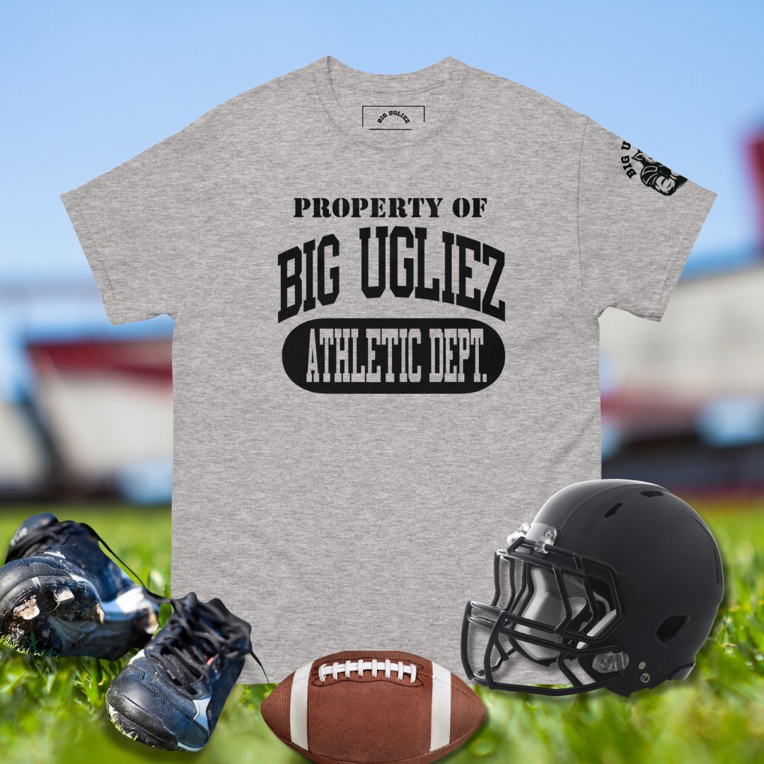 Gray t-shirt with 'Property of Big Ugliez Athletic Dept.' text, black helmet, football, and cleats on grass.