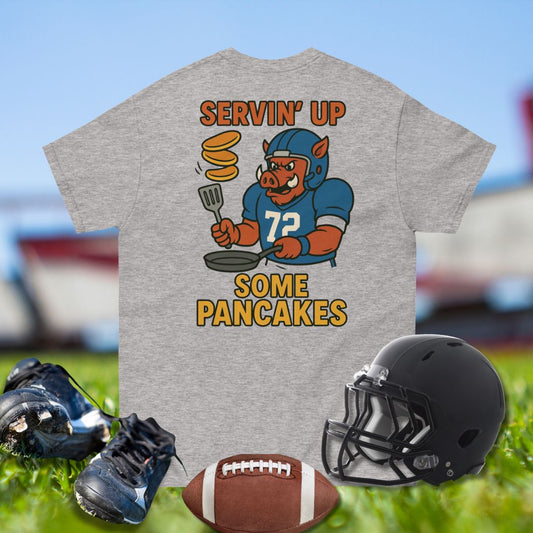 Gray t-shirt with cartoon pig in football uniform and text, surrounded by football helmet, cleats, and ball on grass.