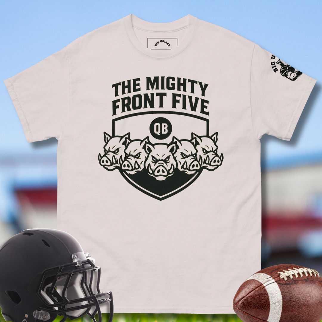 Beige t-shirt with 'The Mighty Front Five' design, black helmet, and football on a blurred sports field background.