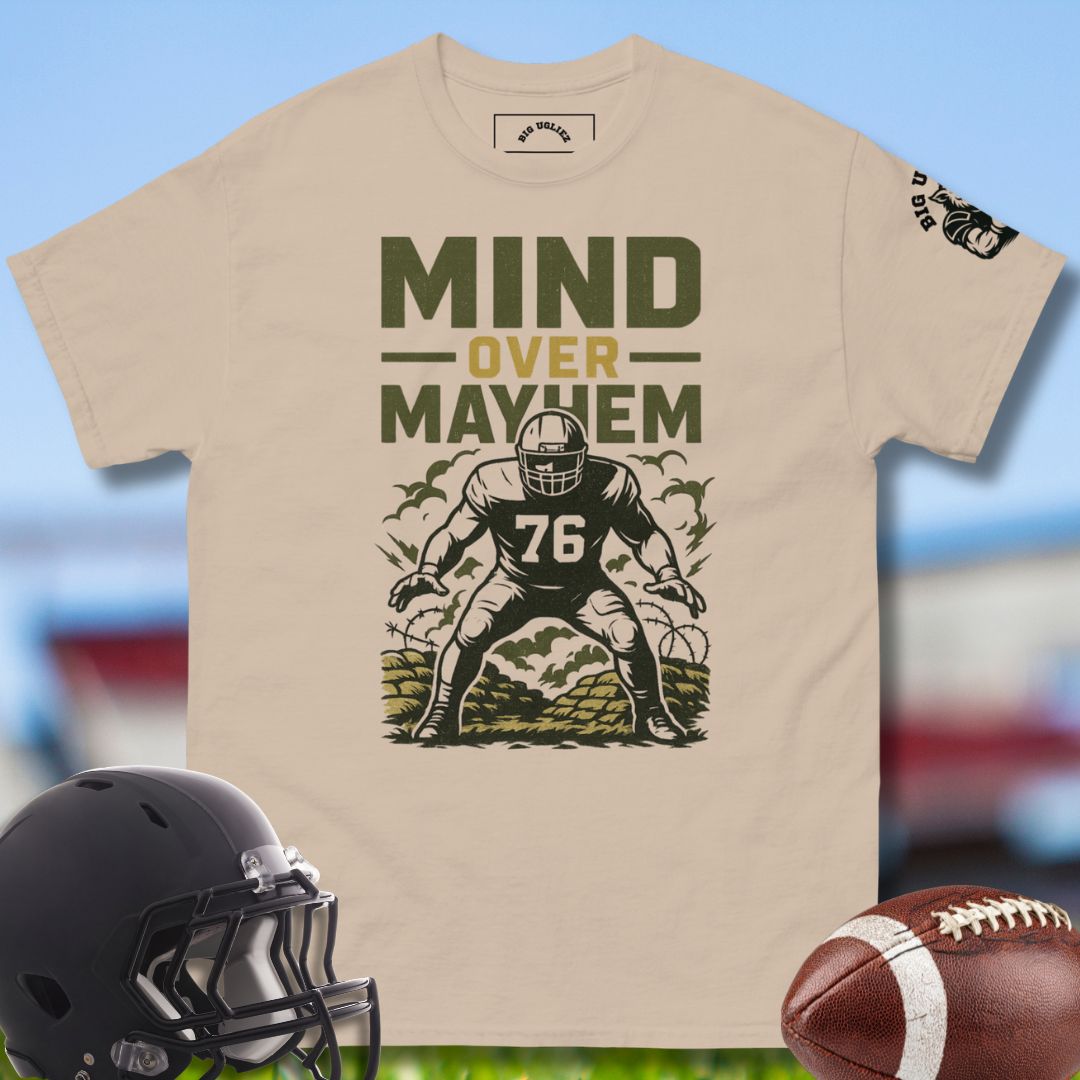 Beige t-shirt with football player graphic and 'Mind Over Mayhem' text, helmet, and football on a blurred sports field background.