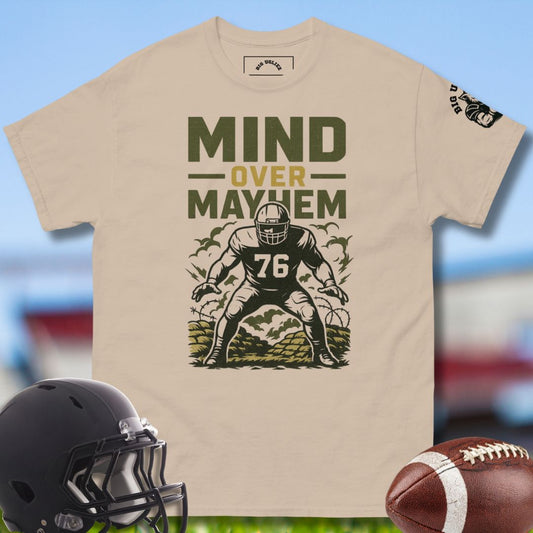 Beige t-shirt with football player graphic and 'Mind Over Mayhem' text, helmet, and football on a blurred sports field background.
