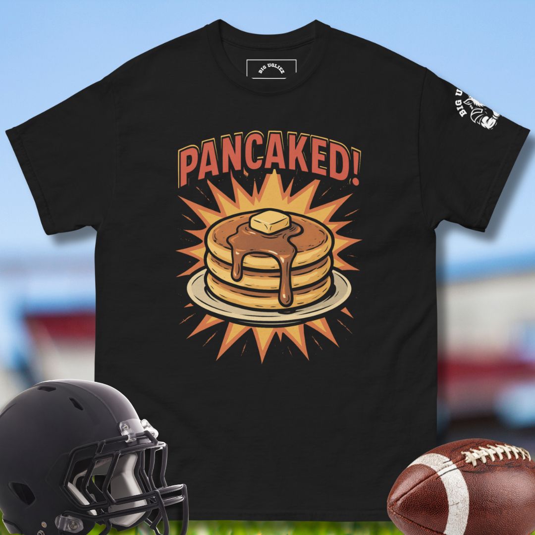 Black t-shirt with 'PANCAKED!' graphic design featuring pancakes and syrup, helmet, and football on a blurred sports field background.