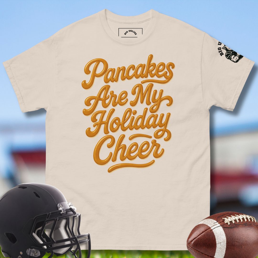 Beige t-shirt with 'Pancakes Are My Holiday Cheer' text, black football helmet, and brown football on a blurred sports field background.