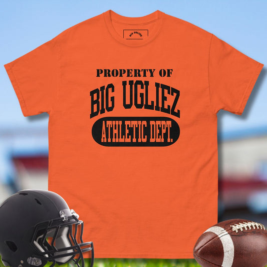 Orange t-shirt with 'Property of Big Ugliez Athletic Dept.' text, black helmet, and football on a blurred sports field background.