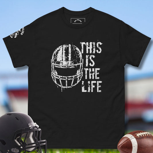 Black t-shirt with football helmet graphic and 'This is the Life' text, displayed with a helmet and football on a sports field background.