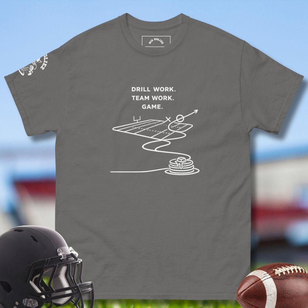 Gray t-shirt with football-themed graphic and text, helmet, and football on a sports field background.