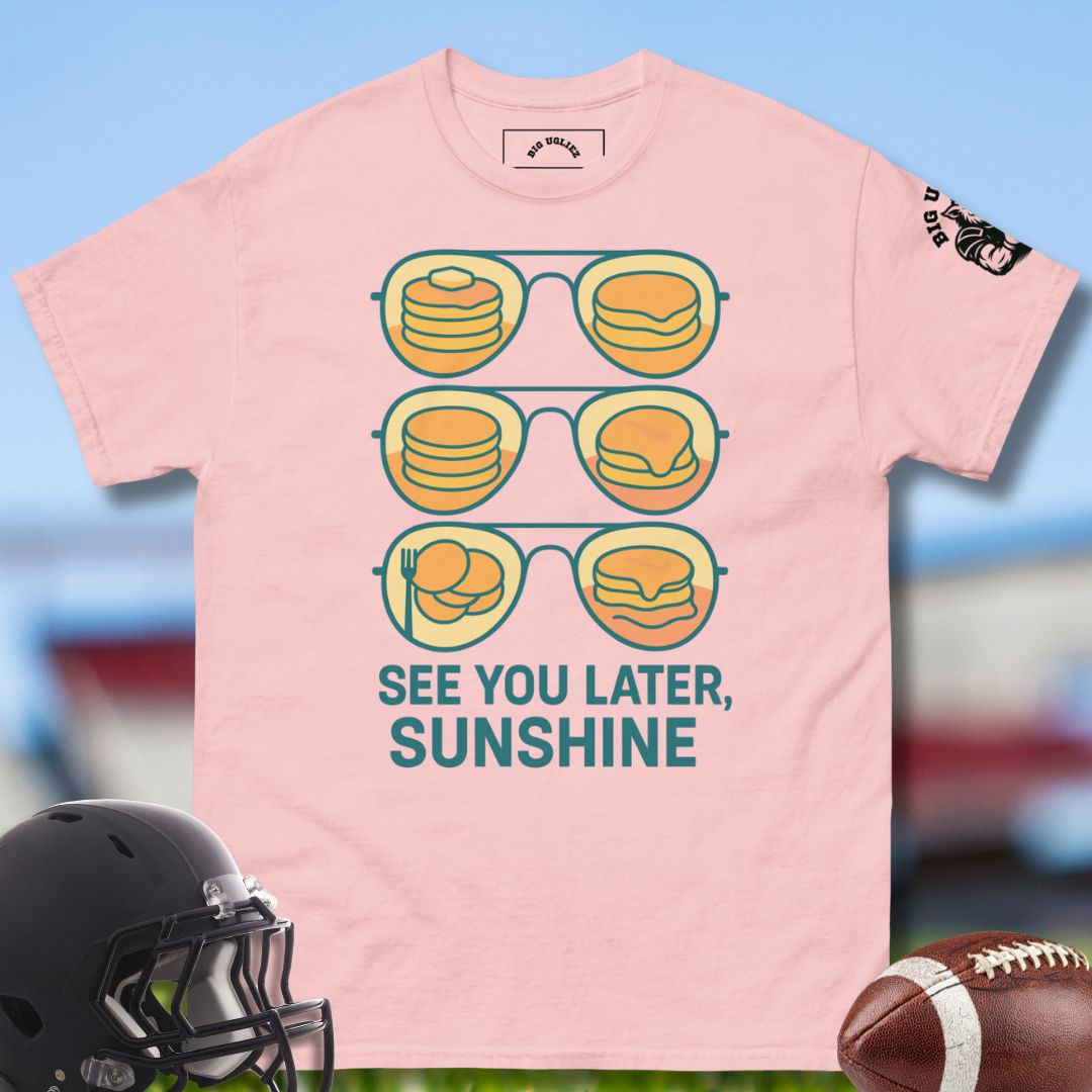 Pink t-shirt with sunglasses and food graphic, text 'See You Later, Sunshine', helmet, and football on a blurred sports field background.