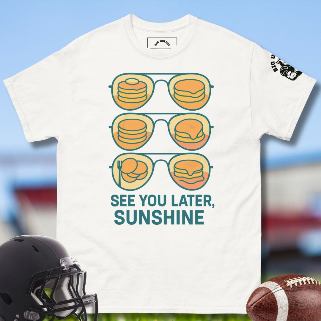 White t-shirt with sunglasses and food graphic and 'See You Later, Sunshine' text, displayed with a football and helmet on a sports field background.